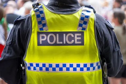 Photograph of the shoulders of a police officer facing with back to the camera. They are wearing a blue short sleeved shirt and fluorescent yellow high vis vest with the word "police" written across it